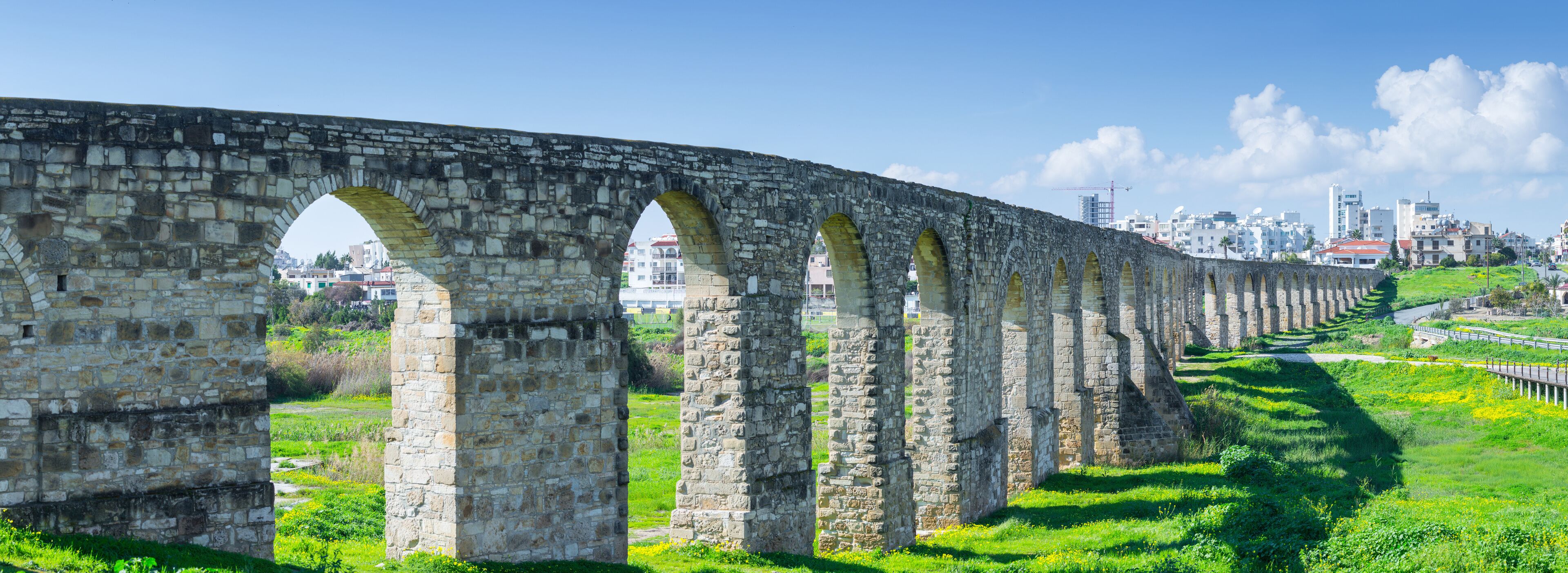 An ancient stone aqueduct with multiple arches, a green landscape, and a cityscape in the background.