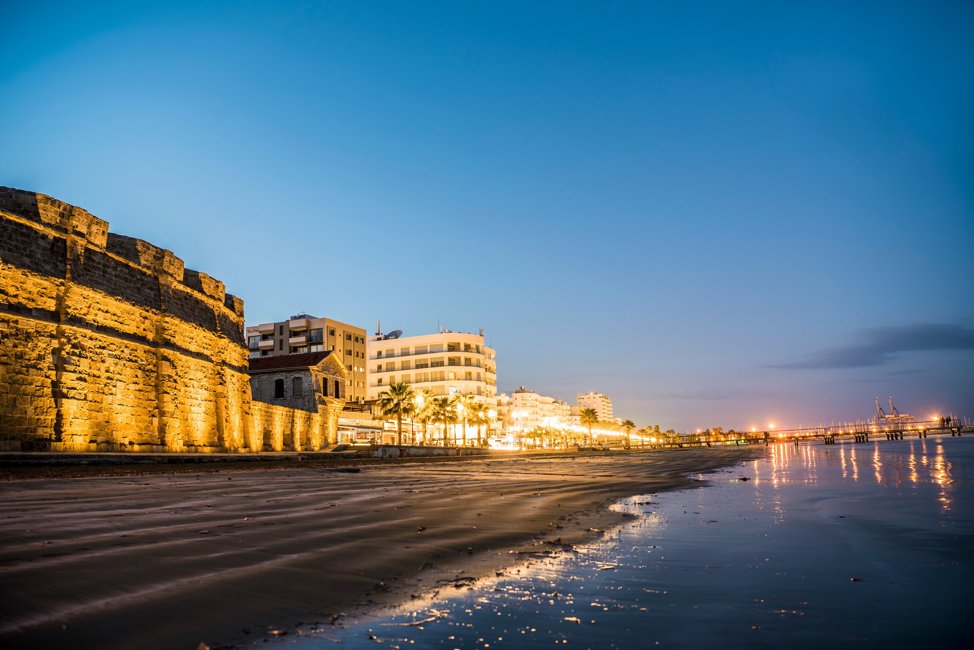 A coastal cityscape at dusk with a beach, a historic stone wall, and modern buildings.