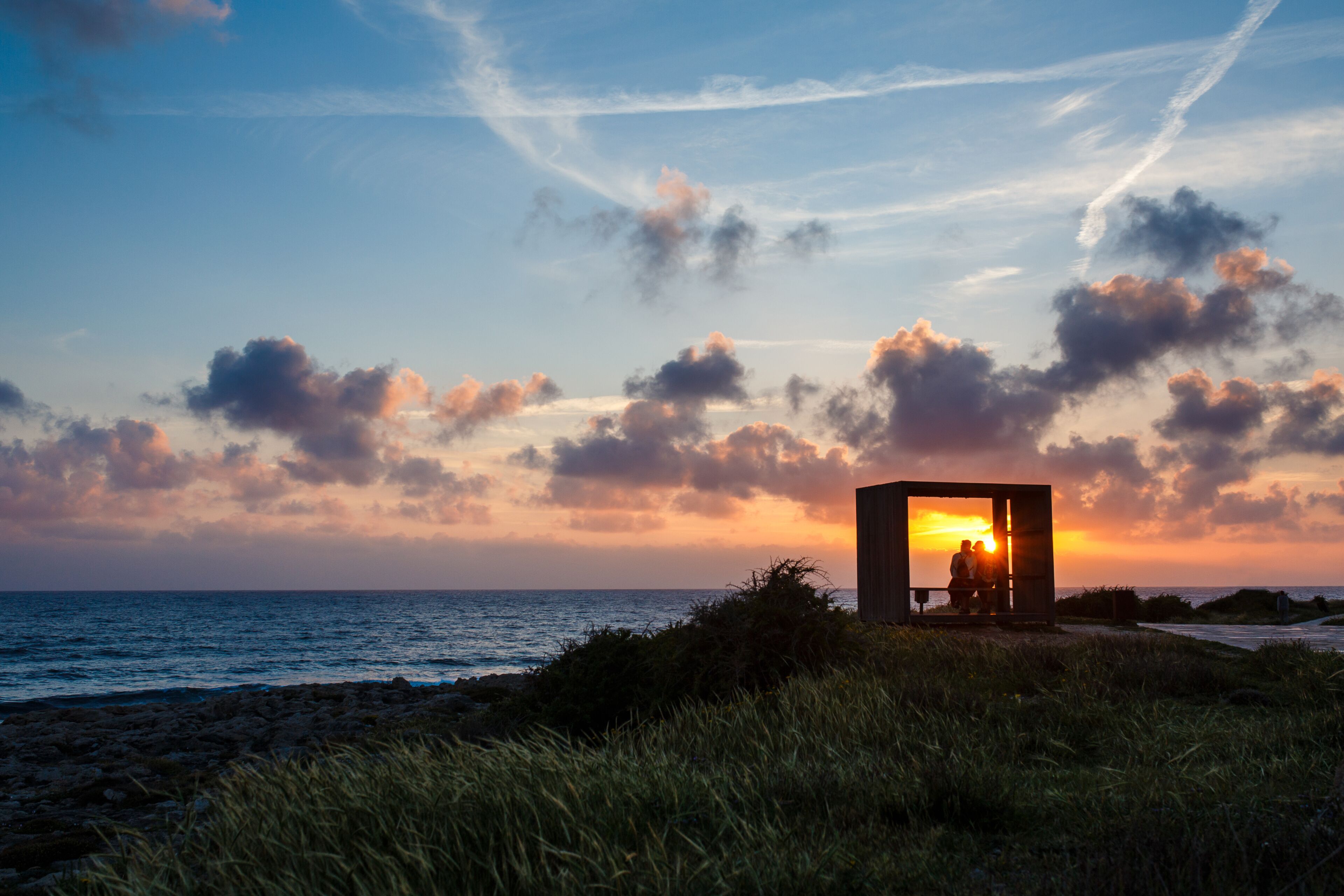 Un atardecer sobre el océano con una estructura de madera y dos personas silueteadas contra el cielo.