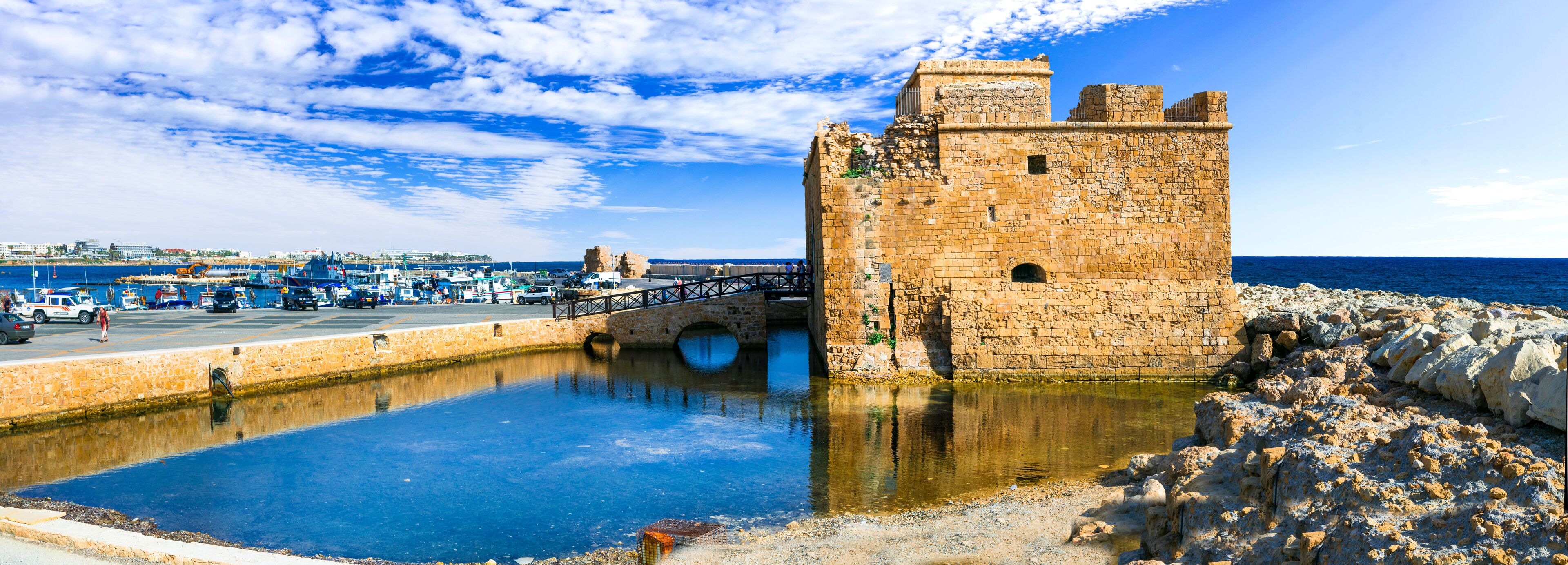 A historic stone tower by a waterfront with a bridge and boats in the background.