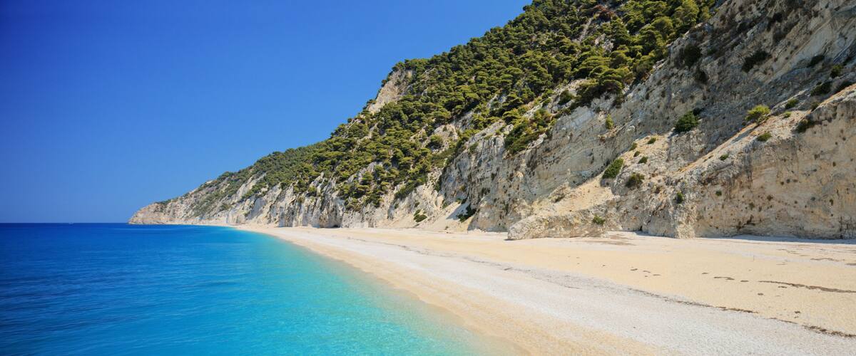 A coastal beach with clear turquoise waters, sandy shore, and a rocky cliff with sparse vegetation.