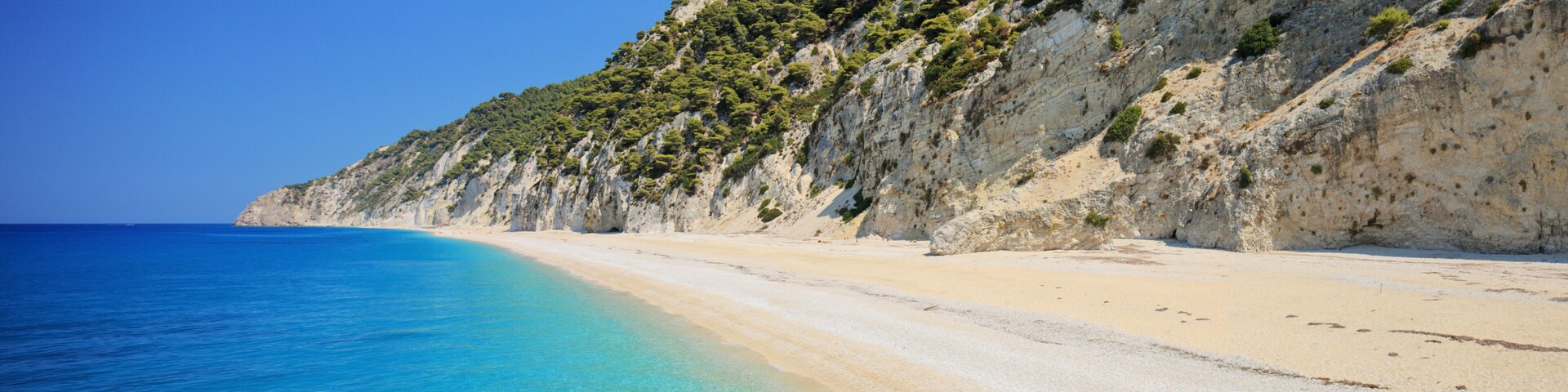 A coastal beach with clear turquoise waters, sandy shore, and a rocky cliff with sparse vegetation.