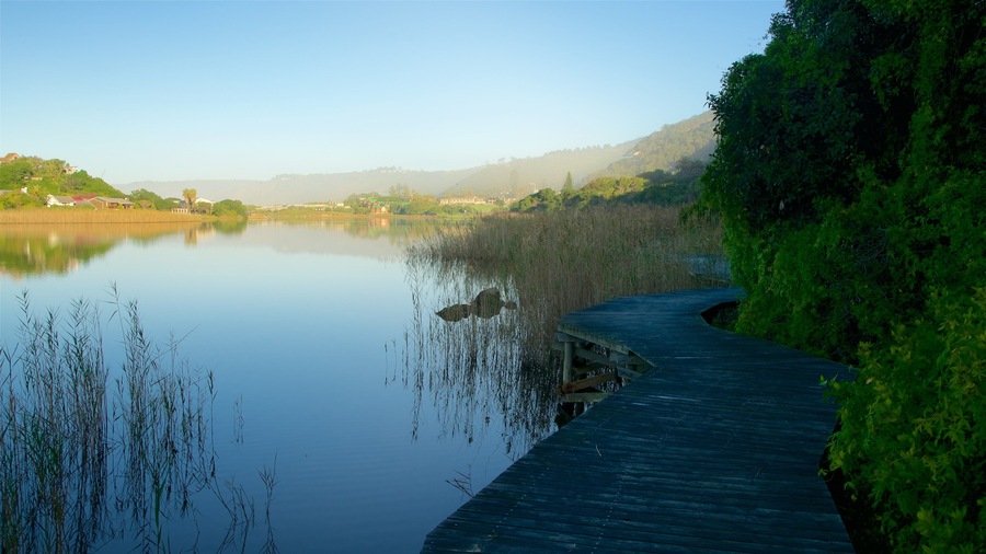 A calm lake with a wooden boardwalk along the right edge, surrounded by dense greenery and hills in the background.
