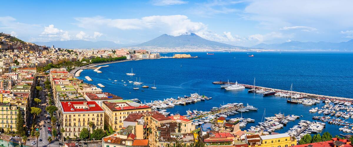 A coastal city with a marina filled with boats, a prominent mountain in the background, and a mix of historic and modern buildings along the waterfront.