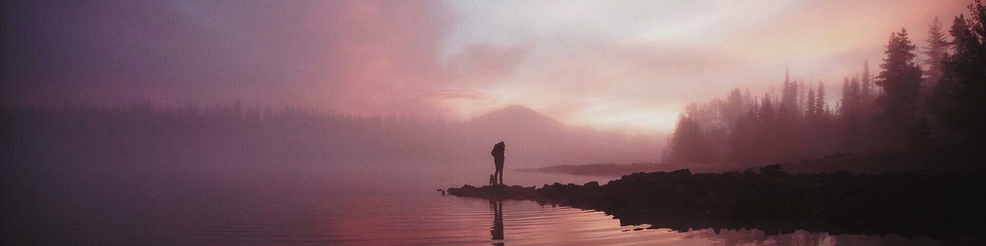 A misty lake with a person standing on a rock, surrounded by trees and a mountain in the background.