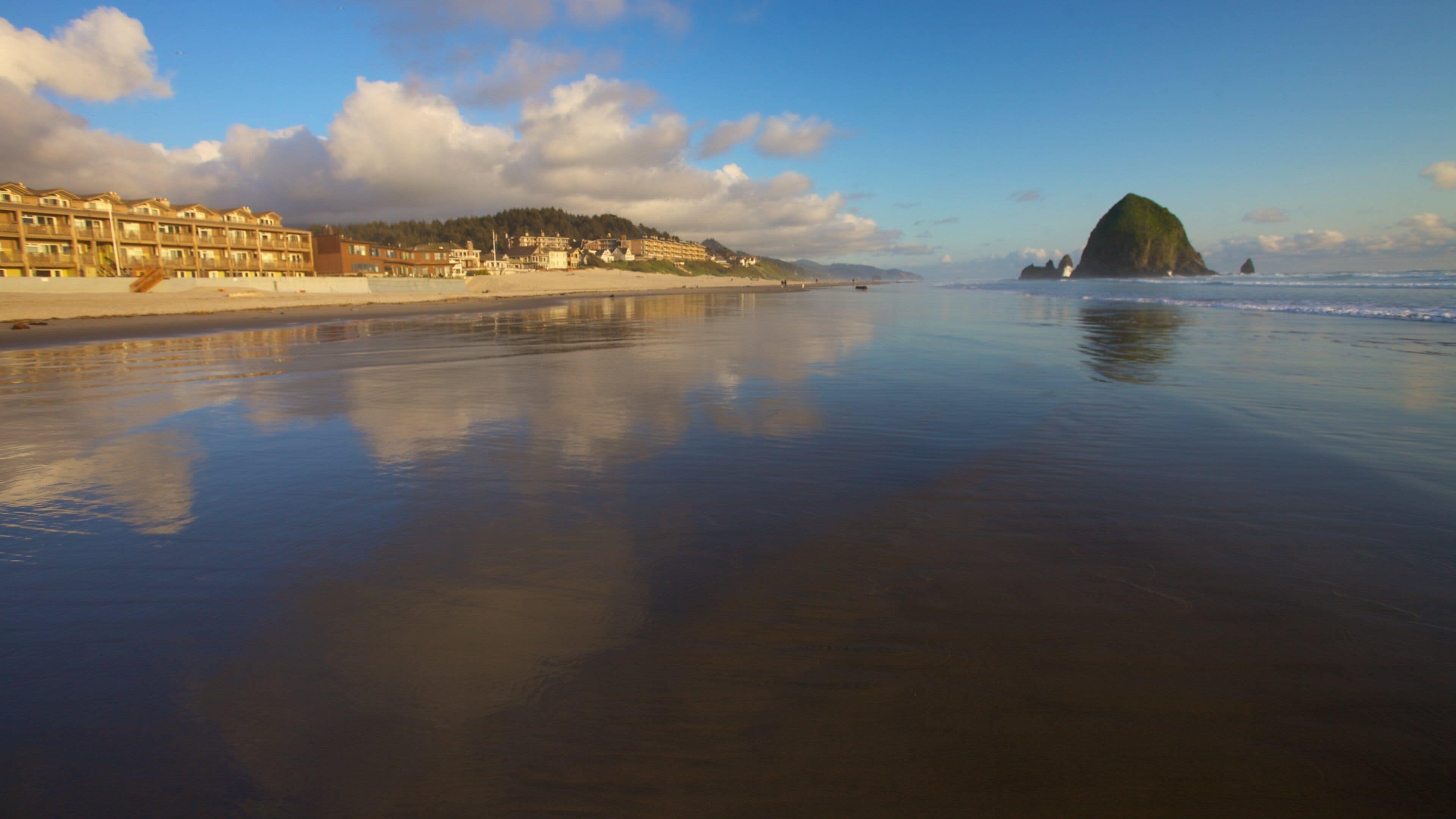 Une plage avec une mer calme, une grande formation rocheuse et des bâtiments qui bordent le rivage.