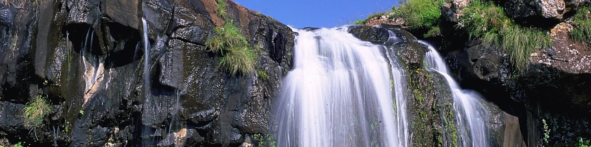 Uma cascata despencando por um penhasco rochoso, cercada por vegetação exuberante.