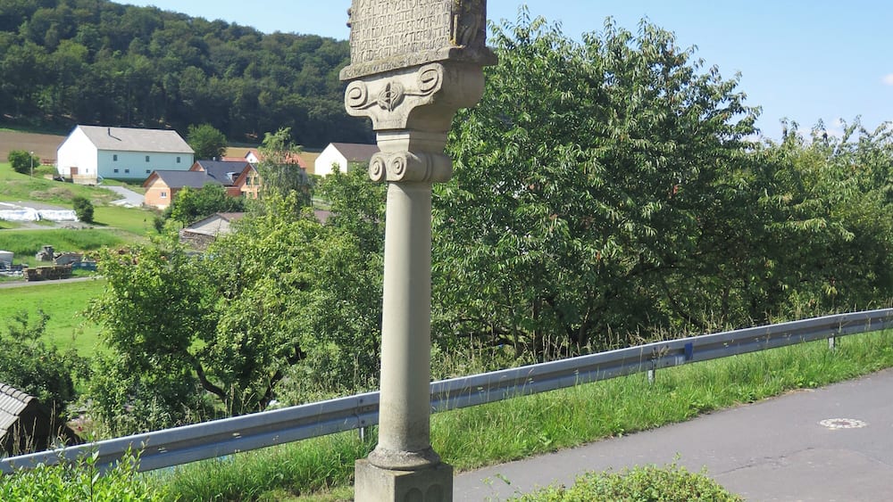 Un monument de pierre surmonté d’une croix, entouré de verdure et bordé par une route.