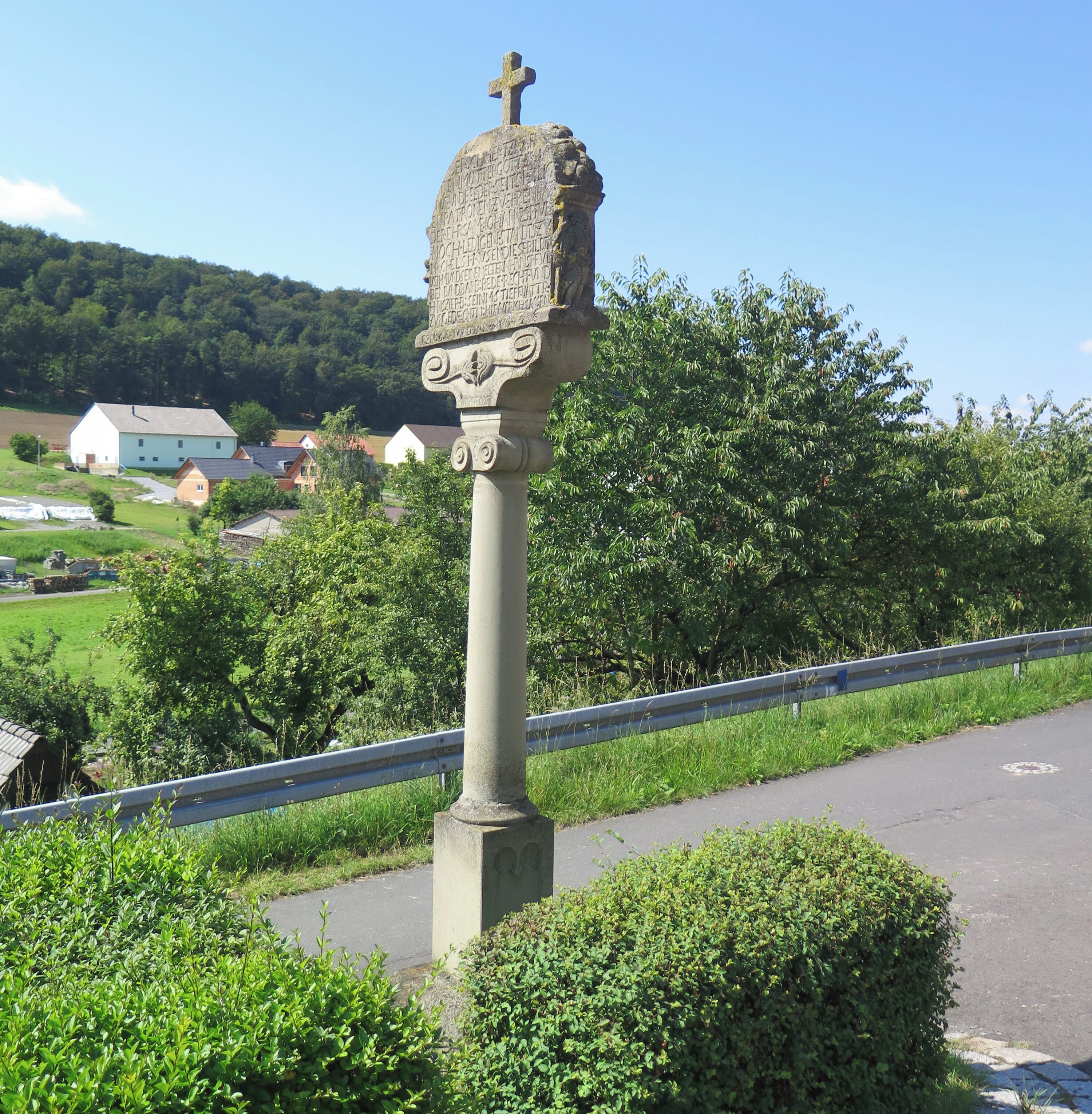 A stone monument with a cross on top, surrounded by greenery and a road.