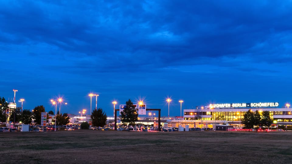 A large airport terminal at dusk with illuminated signs and surrounding infrastructure.