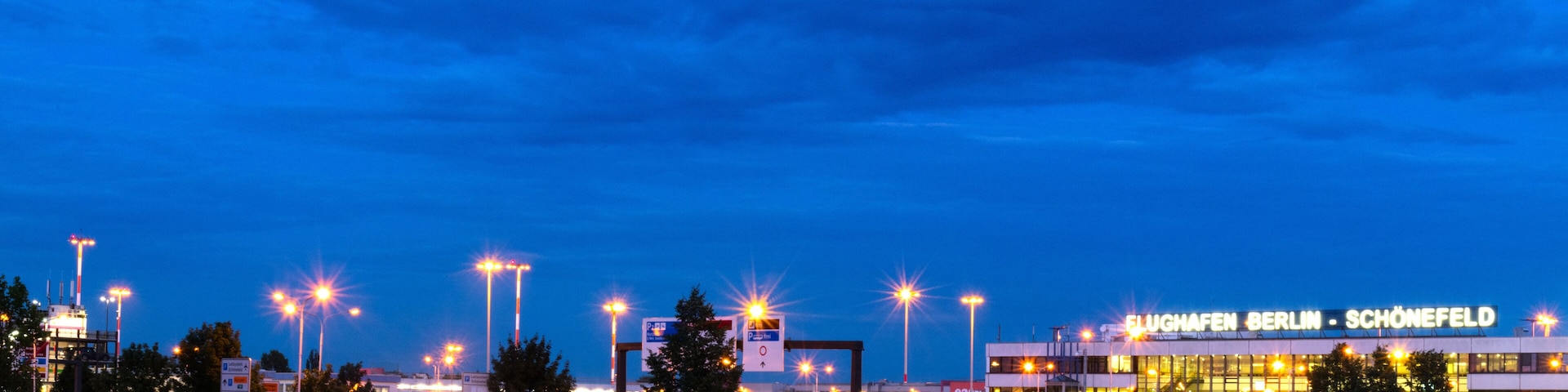 A large airport terminal at dusk with illuminated signs and surrounding infrastructure.