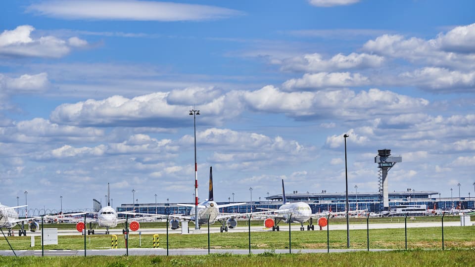 An airport with multiple airplanes on the tarmac, a control tower, and a terminal building in the background.
