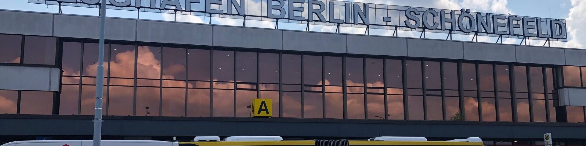 A building with the sign "FLUGHAFFEN BERLIN - SCHÖNEFELD" and a yellow bus in front.
