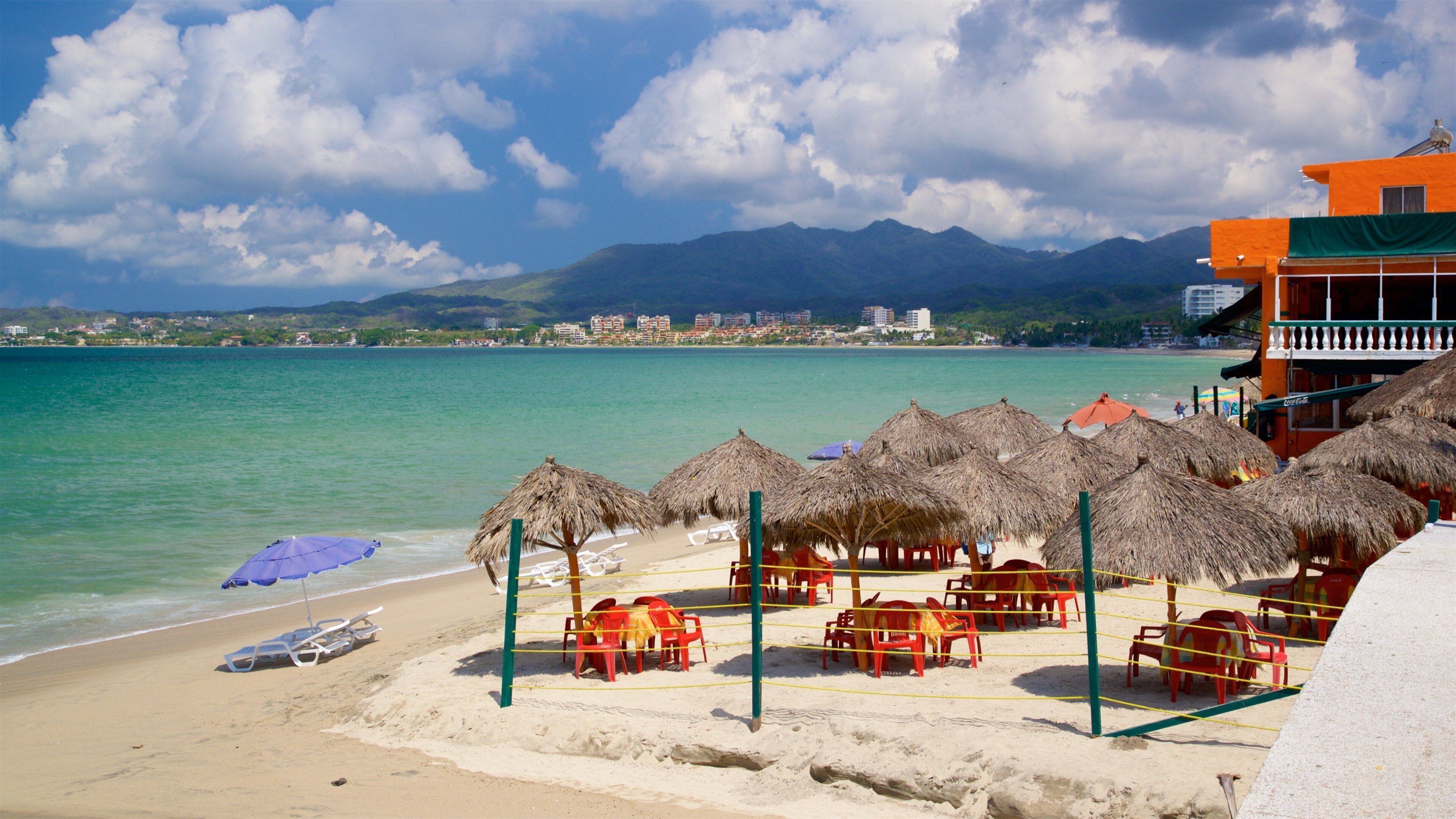 A beach with thatched umbrellas, red chairs, and a clear view of the ocean and mountains in the background.