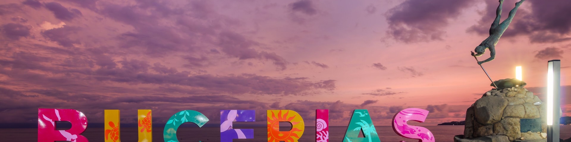Letras coloridas formando la palabra 'BUCERIAS' en una playa con un atardecer de fondo.
