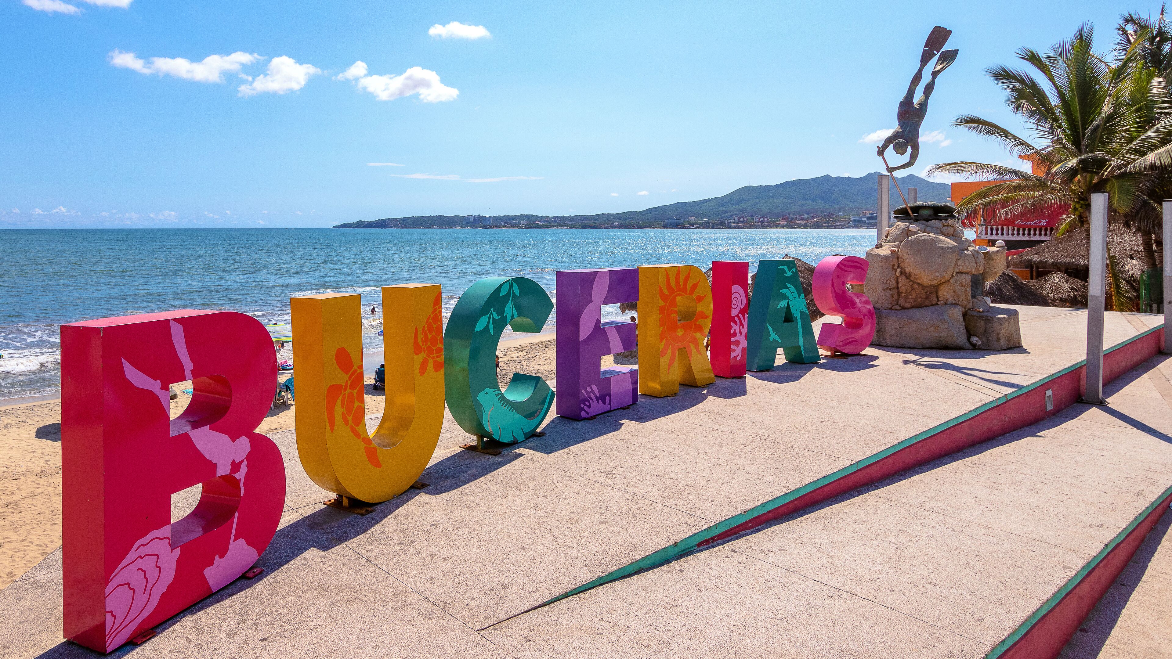Large colorful letters spelling "BUCHERAS" on a beach with the ocean and mountains in the background.