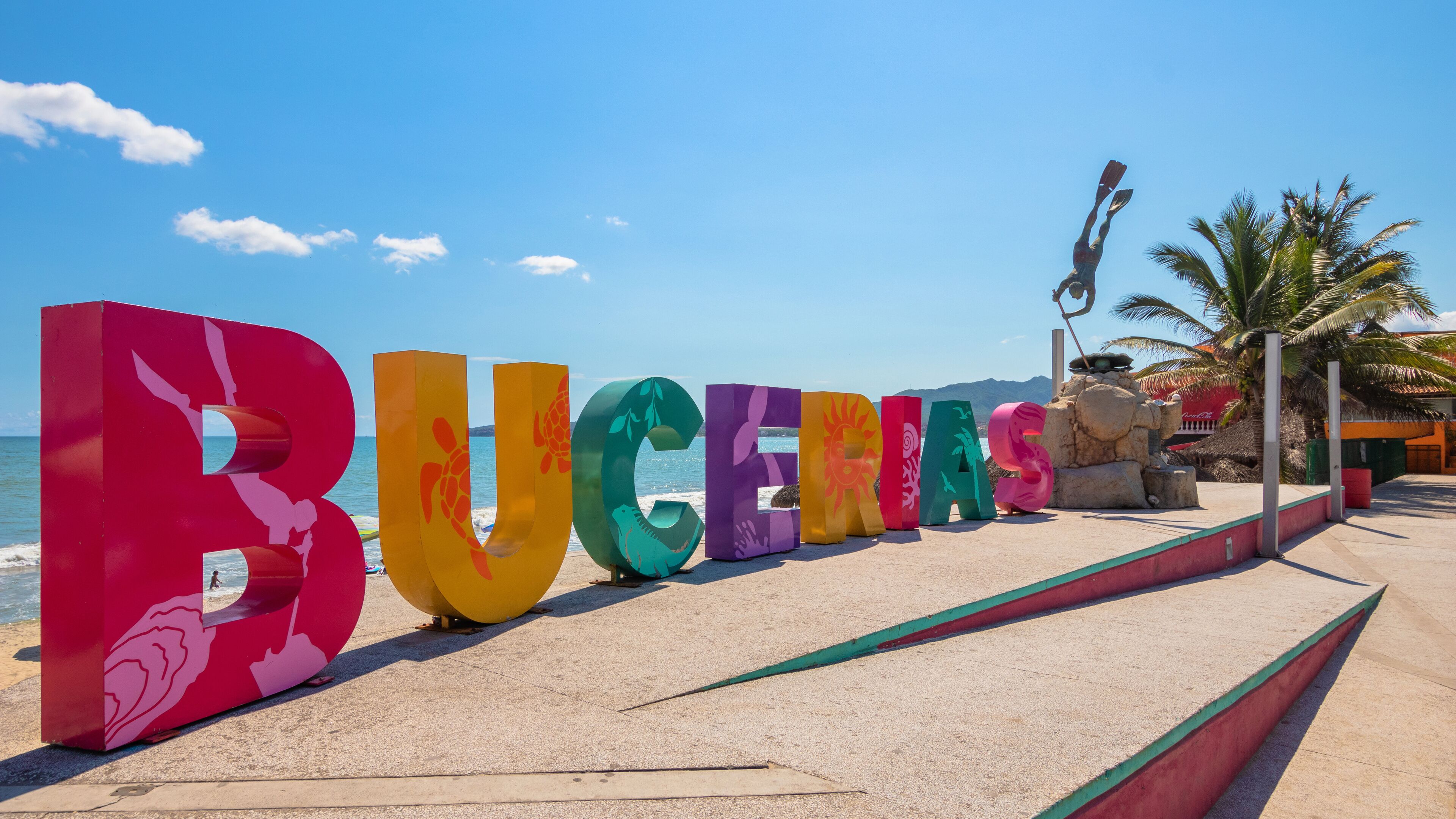 Large colorful letters spelling 'BUCENAS' on a beach with the ocean in the background.