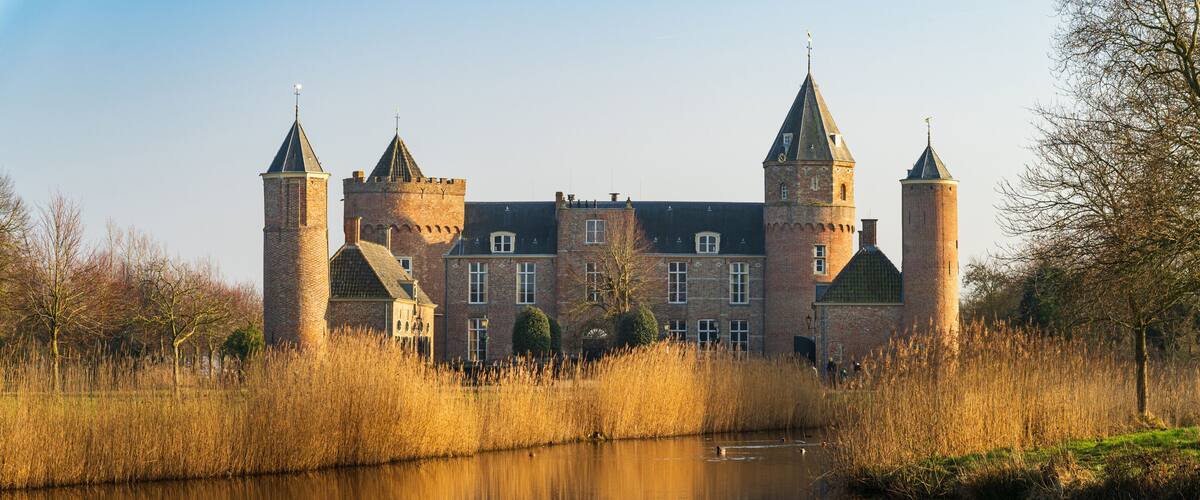 A historic castle with two towers, a moat, and a calm river reflecting the structure.