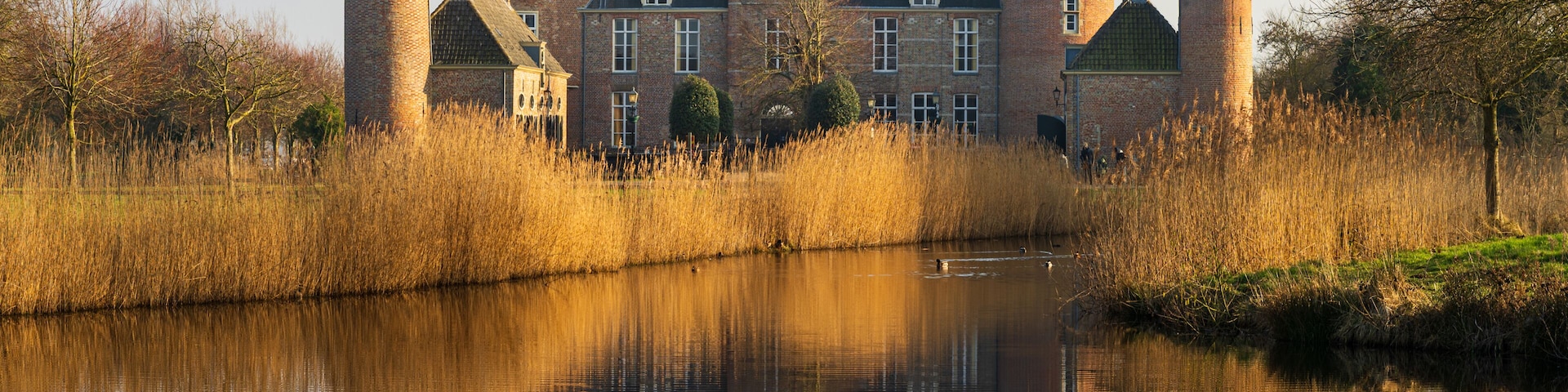 A historic castle with two towers, a moat, and a calm river reflecting the structure.