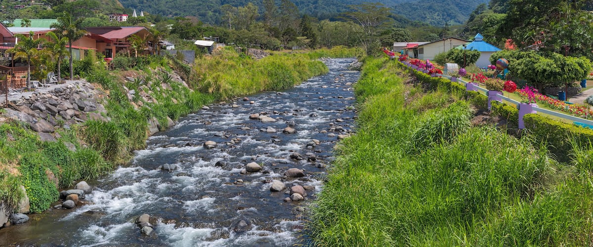 A river flowing through a valley with houses on either side, surrounded by lush greenery and mountains in the background.