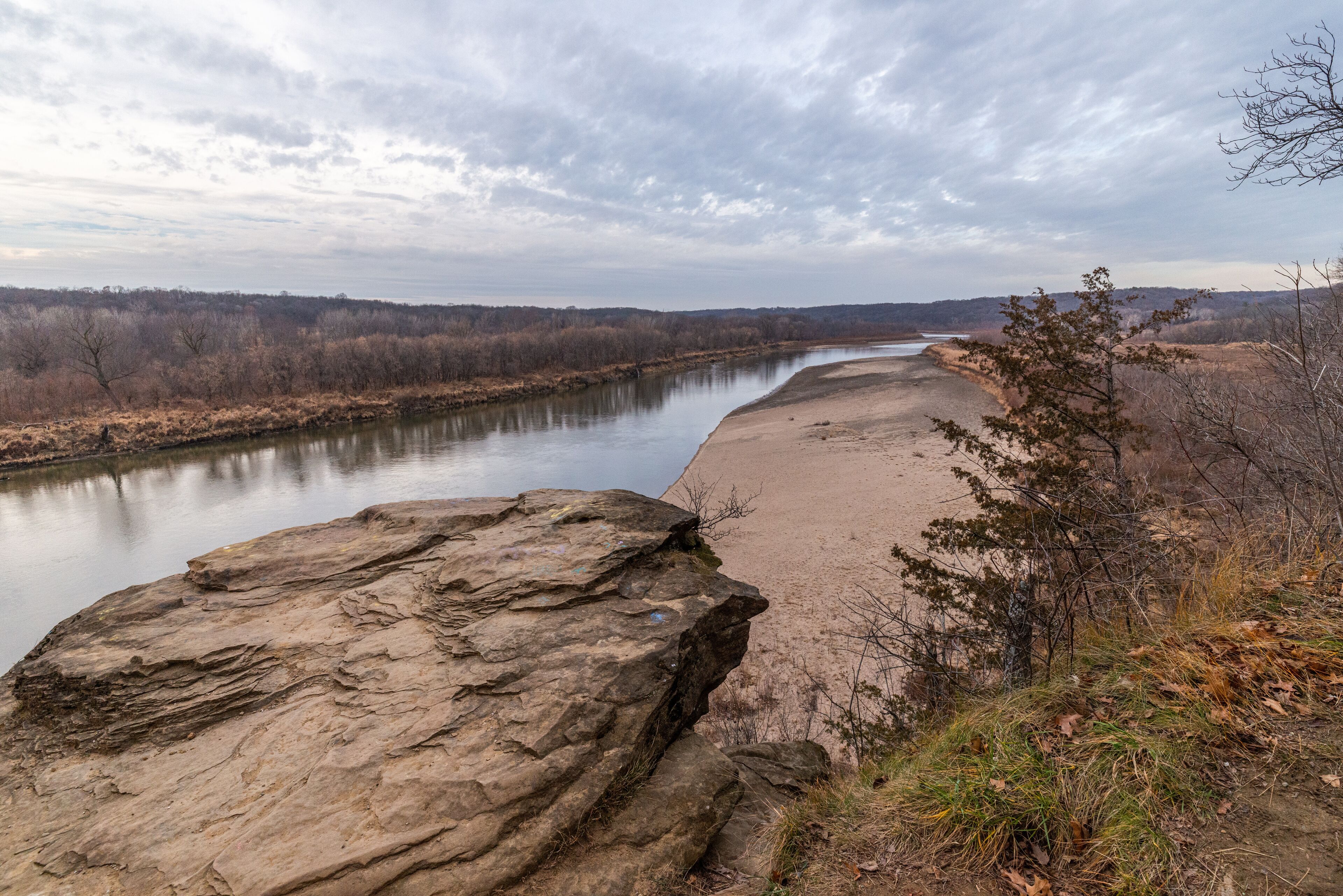 A river with a sandy bank, a large rock in the foreground, and leafless trees in the background.
