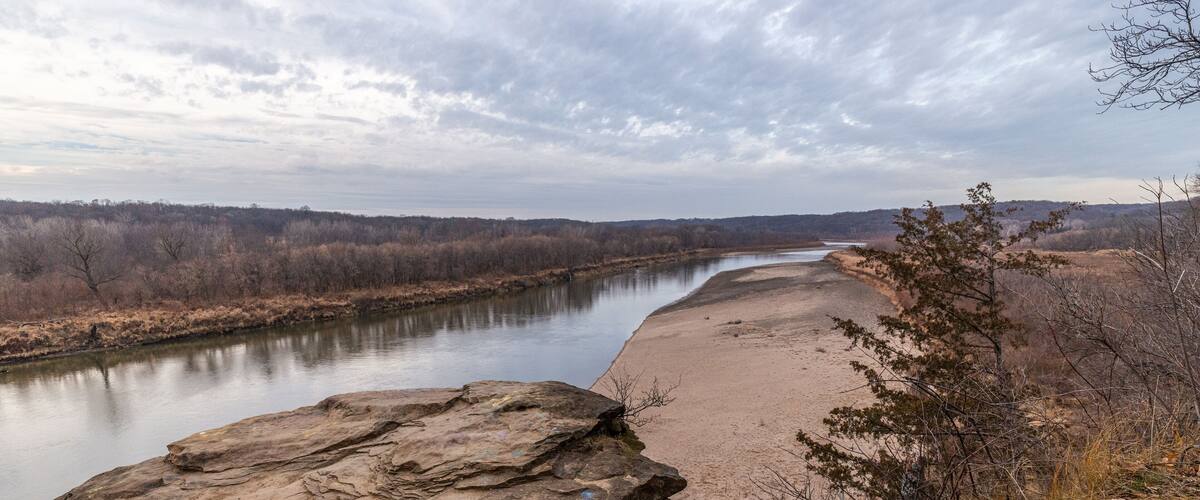 A river with a sandy bank, a large rock in the foreground, and leafless trees in the background.