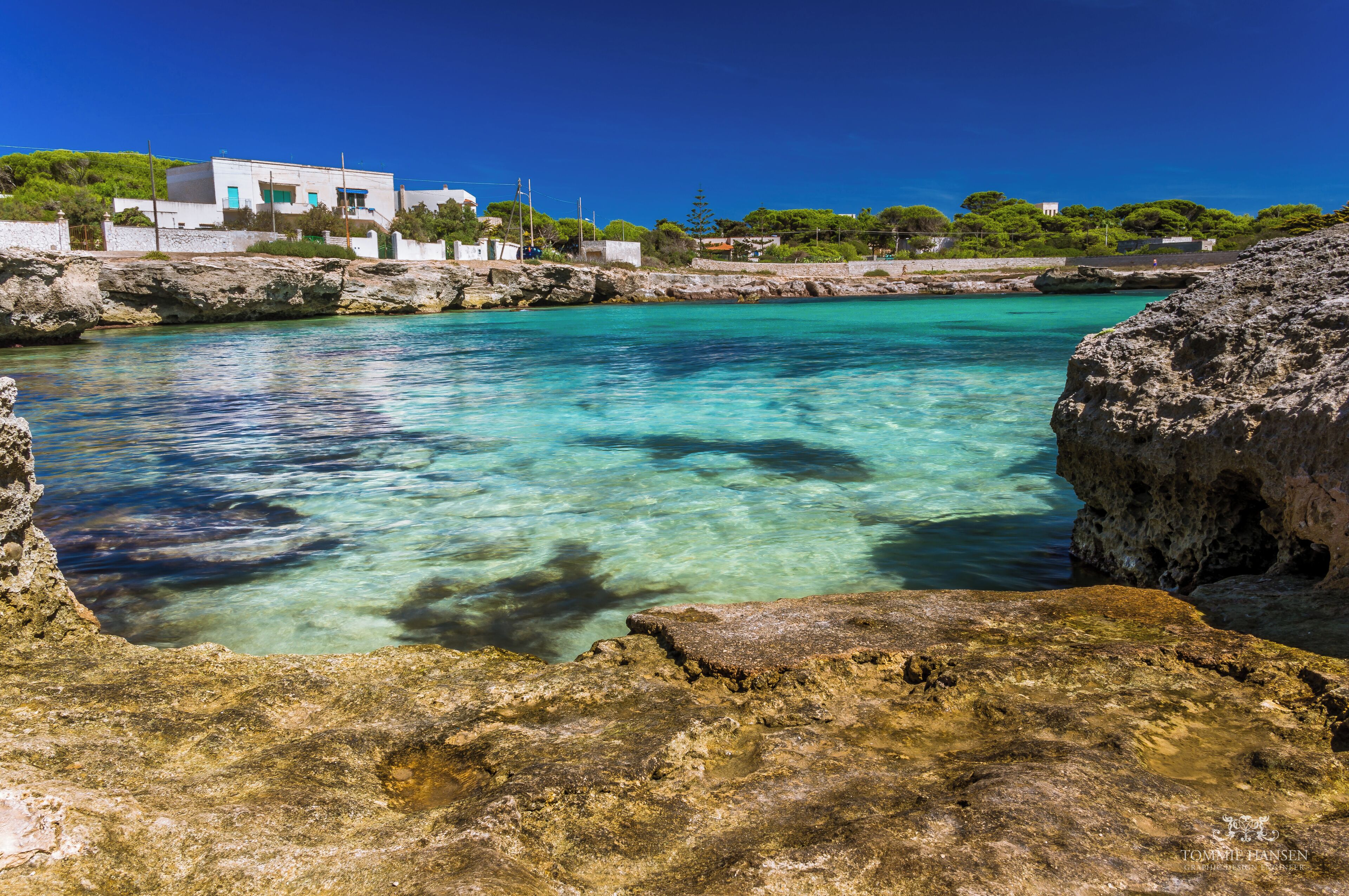A coastal scene with clear turquoise water, rocky formations, and a white building with a blue roof.