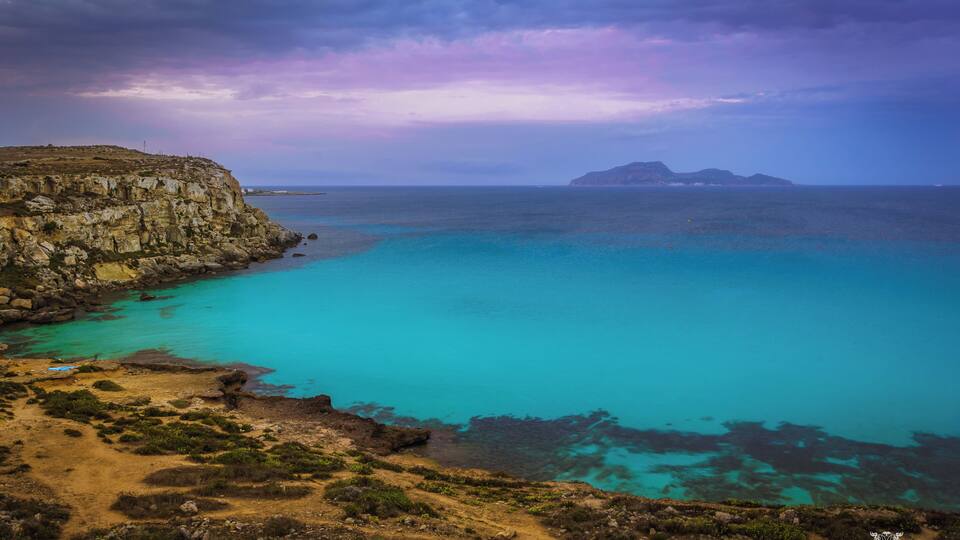 A coastal landscape with a rocky cliff, turquoise waters, and distant islands under a cloudy sky.