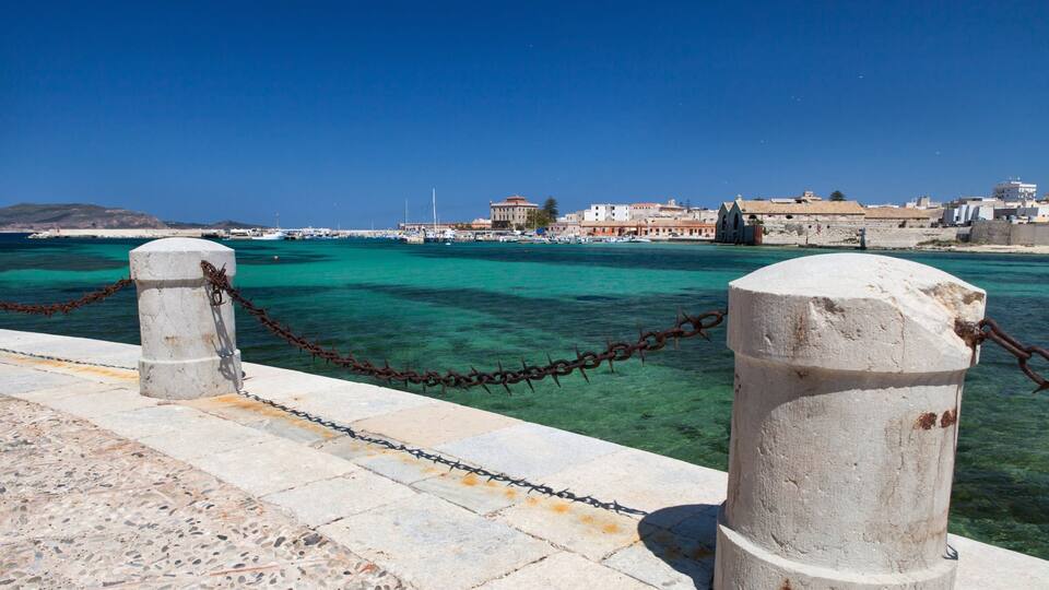 A coastal harbor with clear turquoise waters, a stone pier, and buildings in the background.