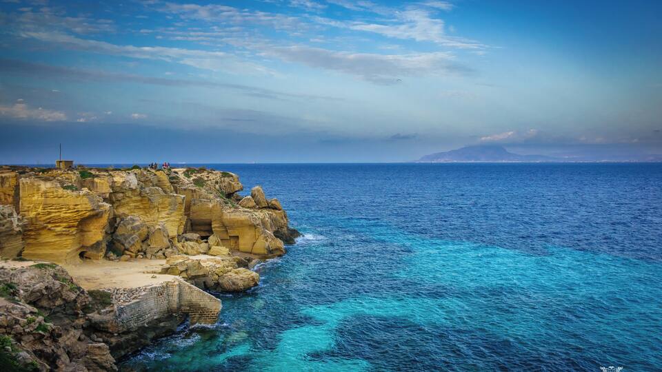 A coastal landscape with rocky cliffs, clear blue sea, and a distant mountain range.