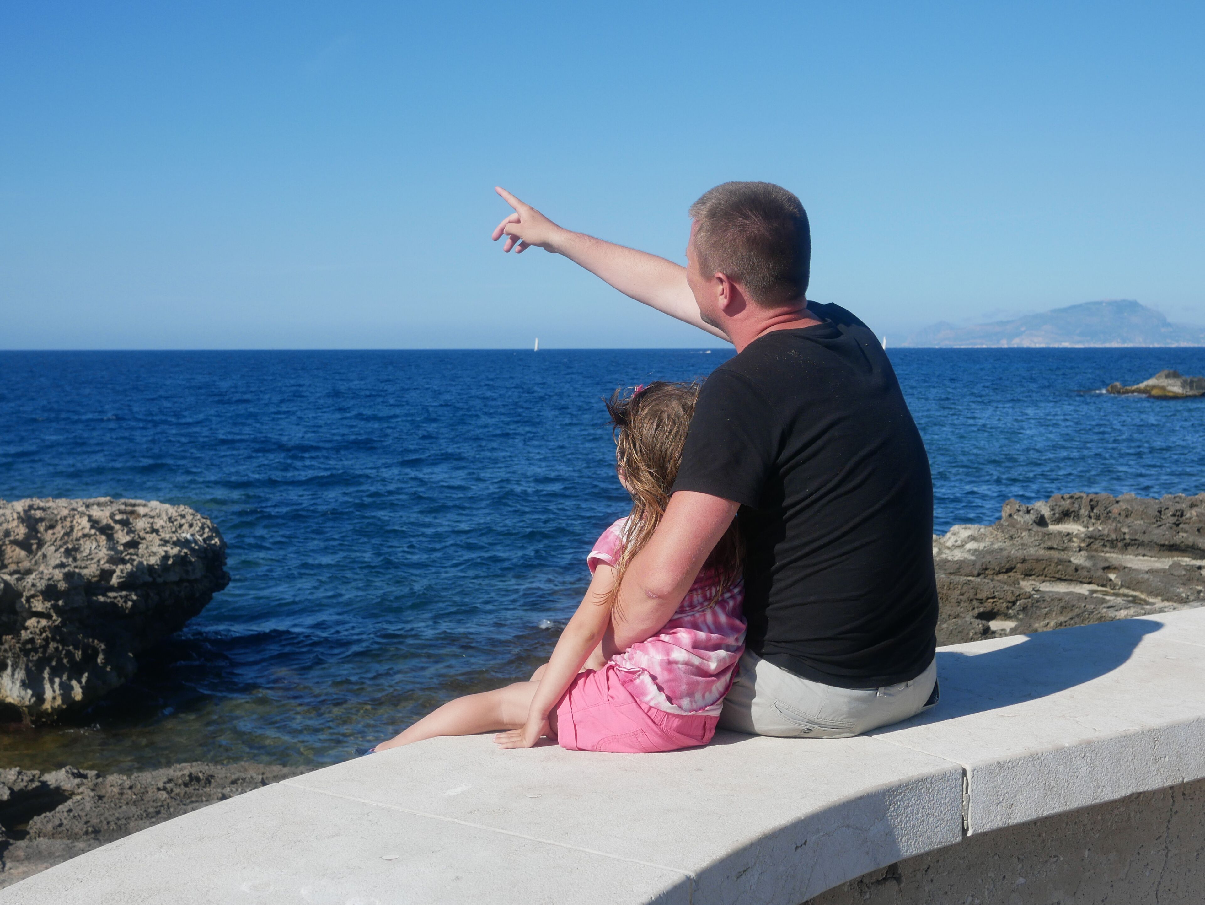 A man and a child sitting on a concrete ledge by the sea, pointing at something in the distance.