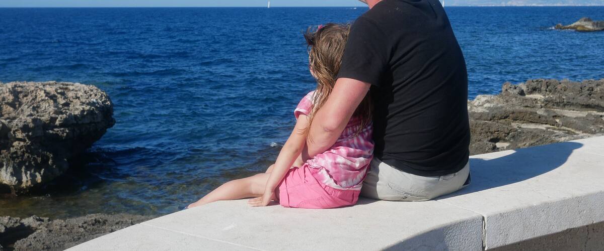 A man and a child sitting on a concrete ledge by the sea, pointing at something in the distance.