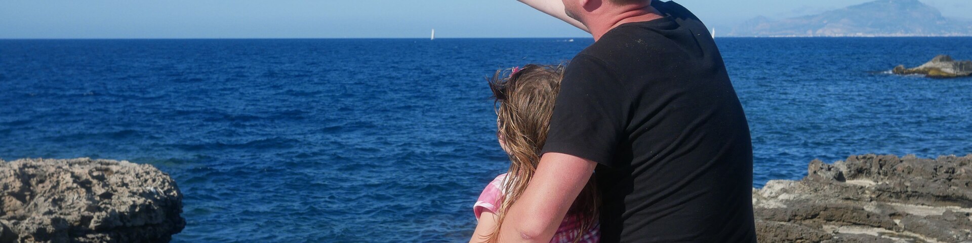 A man and a child sitting on a concrete ledge by the sea, pointing at something in the distance.