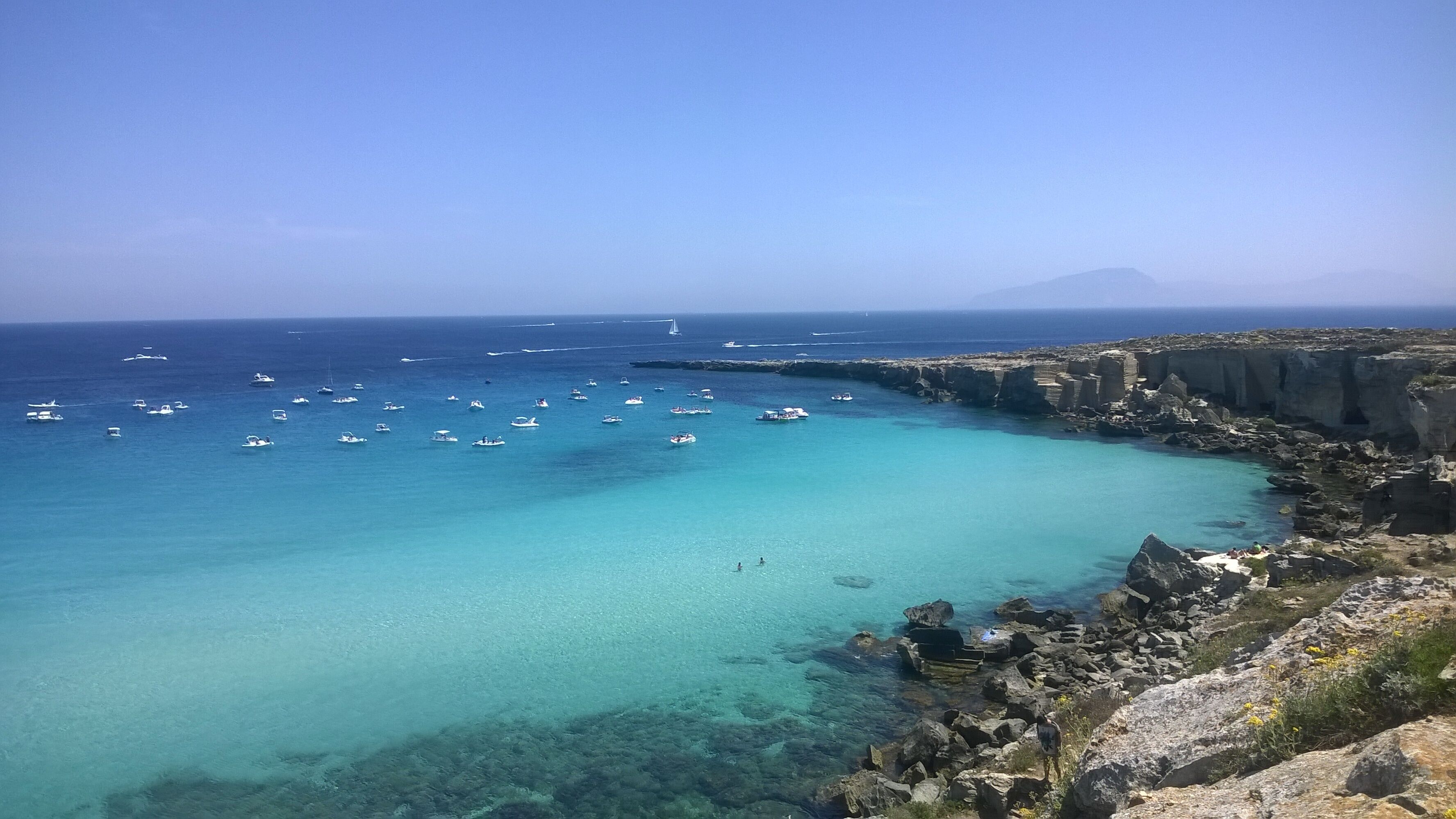 A coastal landscape with clear turquoise waters, rocky cliffs, and numerous boats.