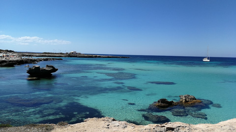 A clear blue sea with a sailboat, rocky shore, and a distant building.