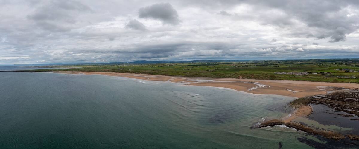 A coastal landscape with a sandy beach, green fields, and a cloudy sky.