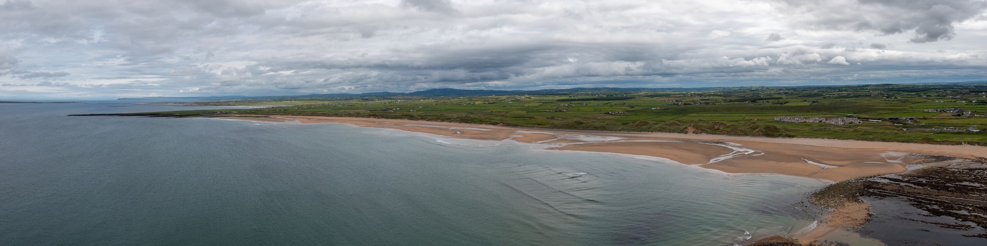 A coastal landscape with a sandy beach, green fields, and a cloudy sky.