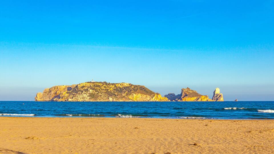 A beach with golden sand, a rocky outcrop, and a lighthouse in the distance.