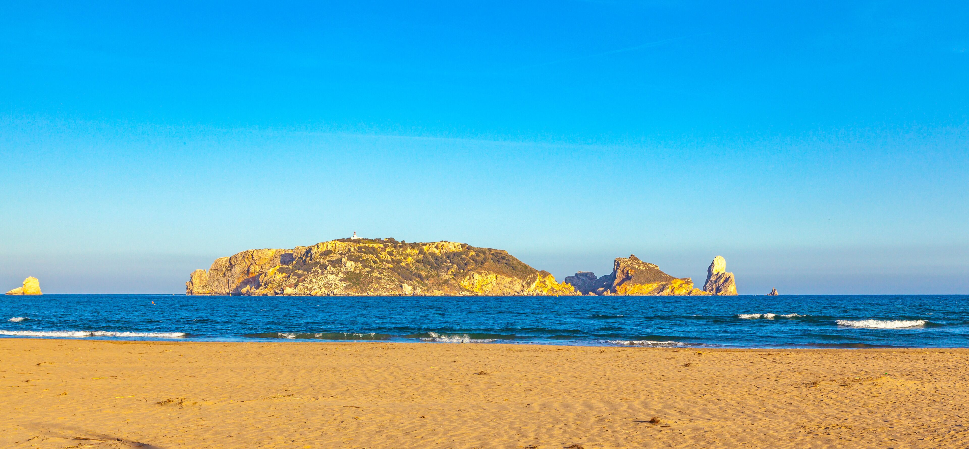 A beach with golden sand, a rocky outcrop, and a lighthouse in the distance.