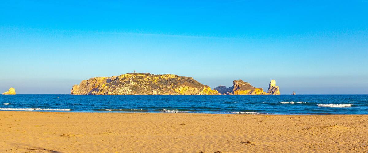 A beach with golden sand, a rocky outcrop, and a lighthouse in the distance.