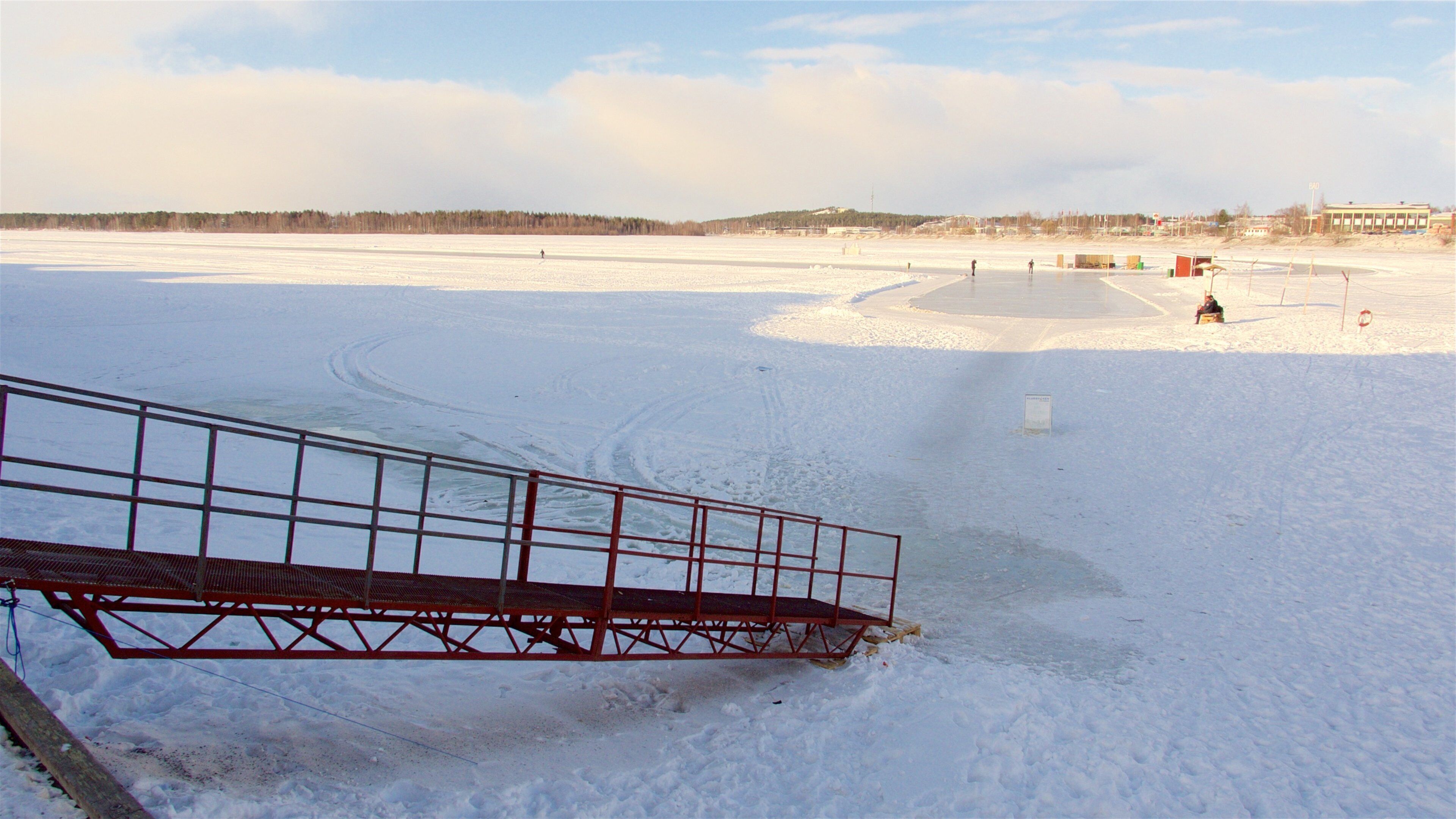 A snowy landscape with a frozen lake, a metal bridge, and distant buildings.