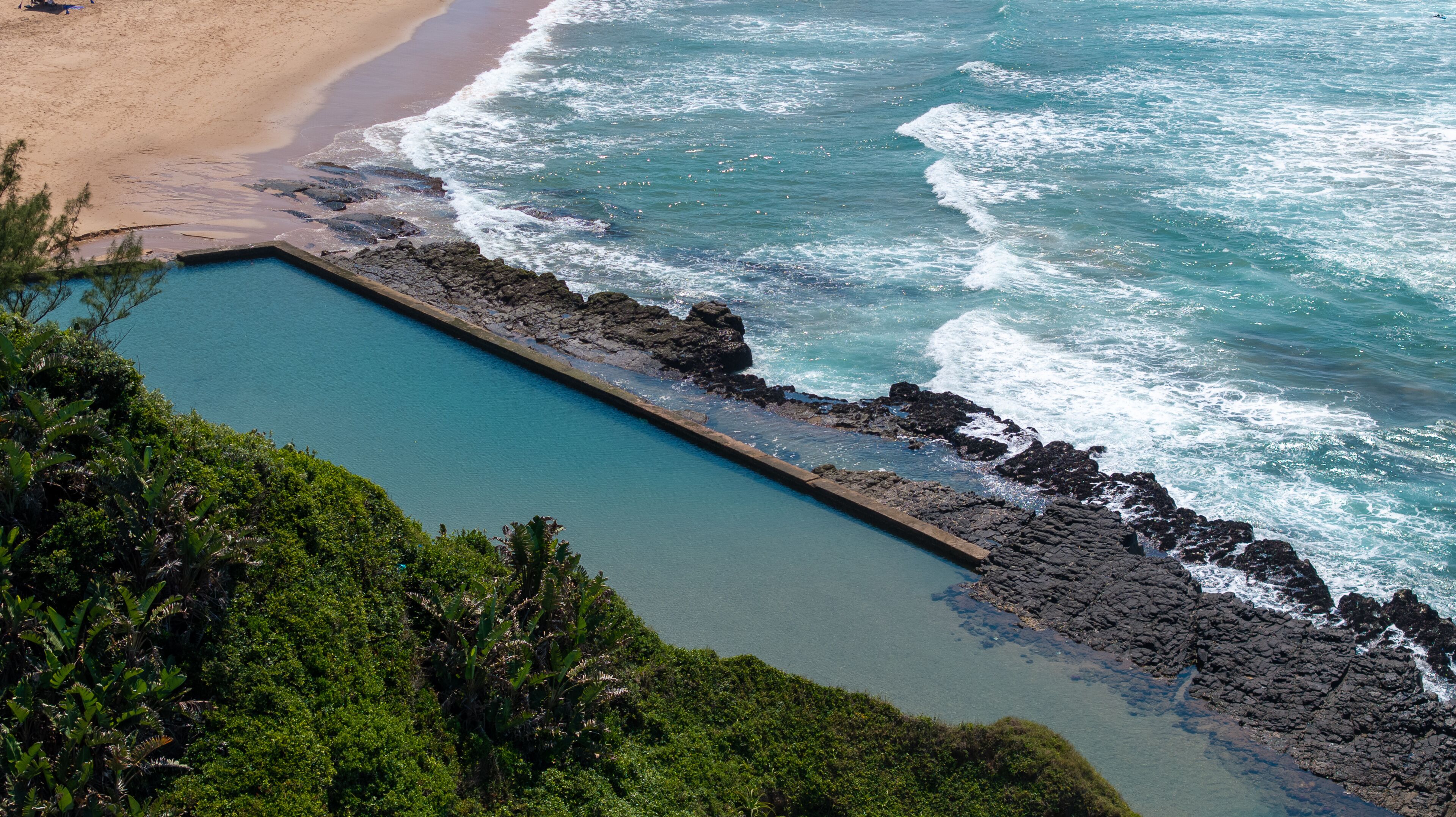 A coastal landscape with a swimming pool, beach, and rocky shoreline.