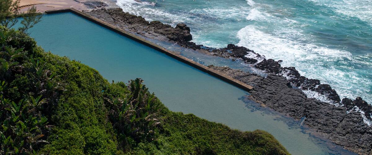 A coastal landscape with a swimming pool, beach, and rocky shoreline.