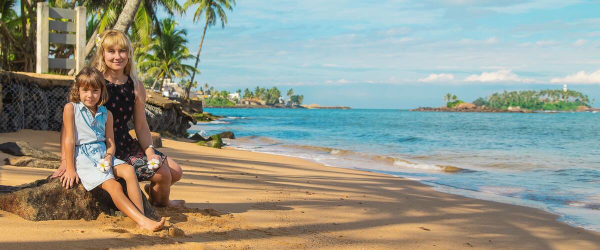 Two people sitting on a sandy beach with palm trees and a calm sea in the background.