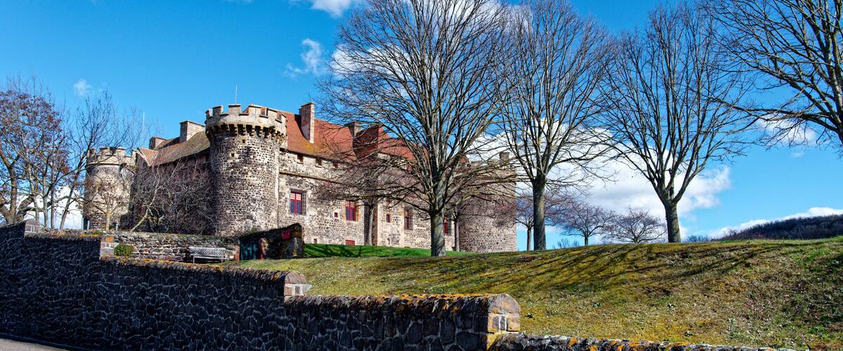 A historic stone castle with a red-tiled roof, surrounded by bare trees and a stone wall.