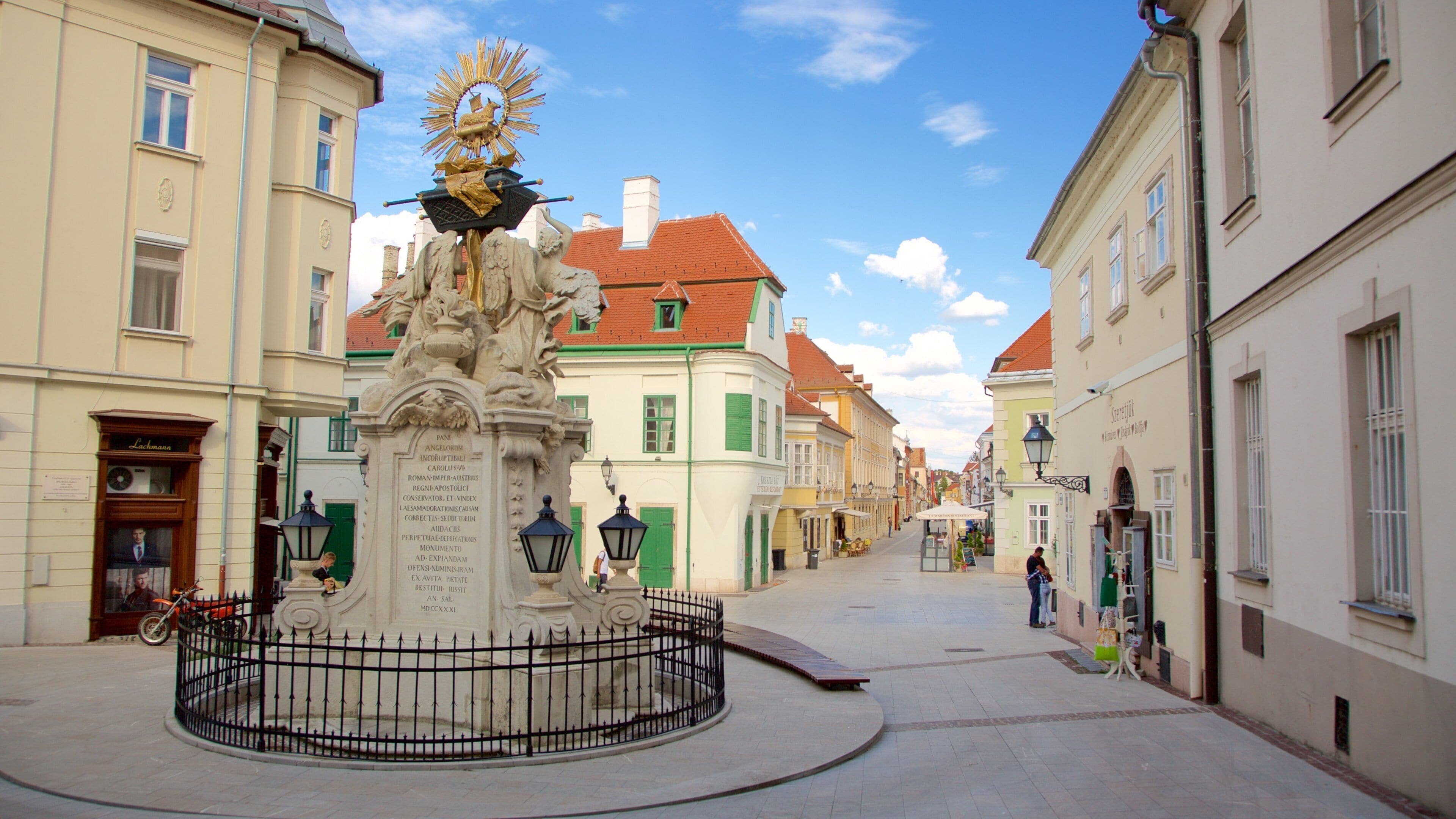 Una plaza histórica con un monumento central, rodeada de edificios europeos tradicionales con tejados de tejas rojas.
