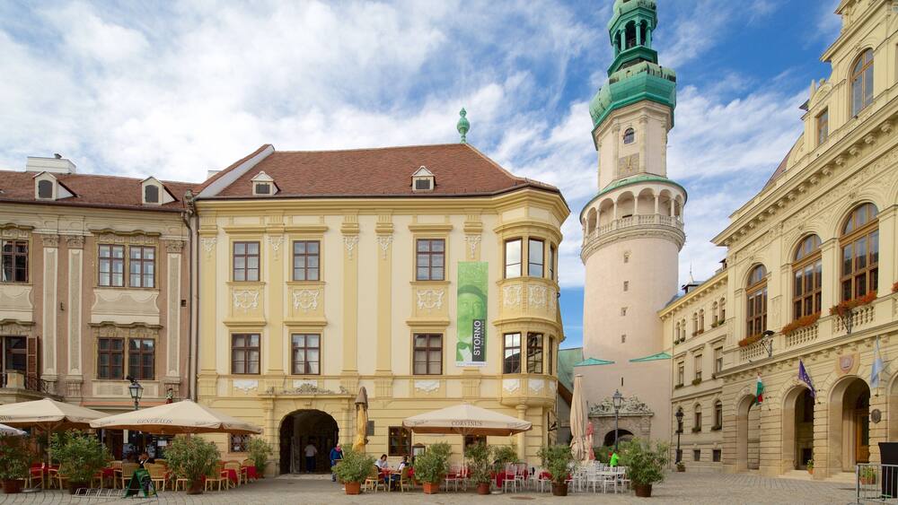 A historic square with a prominent tower, a café area with outdoor seating, and ornate architecture.