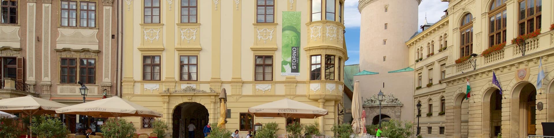 A historic square with a prominent tower, a café area with outdoor seating, and ornate architecture.