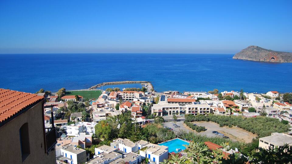 A coastal town with white buildings, a blue pool, and a rocky island in the distance.