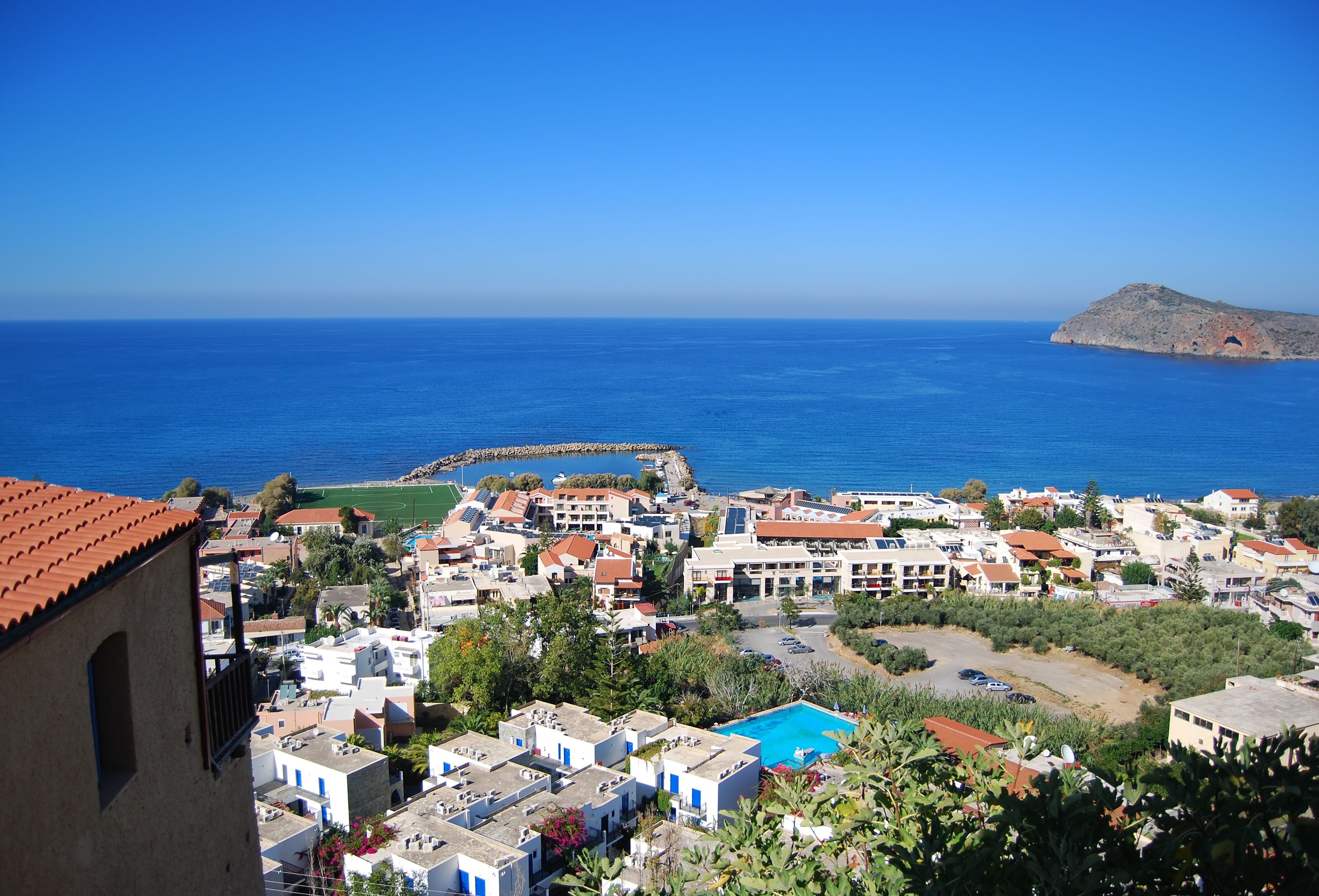 A coastal town with white buildings, a blue pool, and a rocky island in the distance.